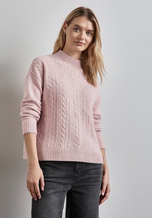 Lichtroze kabelgebreide trui met een hoge ribbels ronde hals en lange mouwen. De stof lijkt zacht en gestructureerd, wat goed aansluit bij donkere jeans.