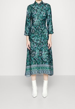 Vrouw draagt een groene midi-jurk met paisley-patroon, lange mouwen, een kraag en witte enkellaarzen, staand tegen een effen achtergrond.