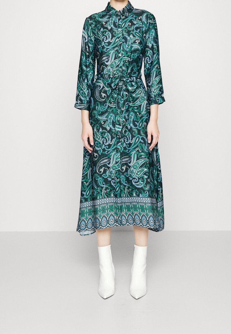 Femme portant une robe midi verte à motif cachemire, manches longues, col, et des bottines blanches, debout devant un fond uni.