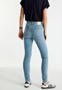 Jeans skinny de mezclilla azul claro con cintura alta, bolsillos traseros y una etiqueta marrón en la cinturilla, combinados con zapatillas negras y blancas.