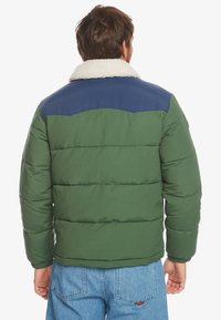 Grøn pufferjakke med marineblå skulderpanel og creme fleecekrave, med vandret quiltning og ribbede manchetter. Casual design.
