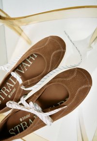 Ballerines en daim marron avec lacets blancs noués en nœud, coutures visibles, et branding "STRADIVARI" sur les semelles intérieures, posées sur une chaise transparente.