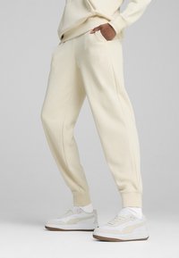 Crème-kleurige sweatpants met een gladde textuur, voorzien van zijzakken, geribde boorden en een geborduurd logo. Gecombineerd met witte sneakers.
