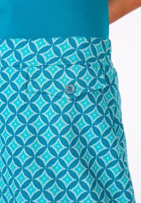 Türkisfarbener Rock mit geometrischen Mustern, ausgestattet mit einer knöpfbaren Tasche und glatter Textur, ergänzt durch ein einfarbiges blaues Oberteil.