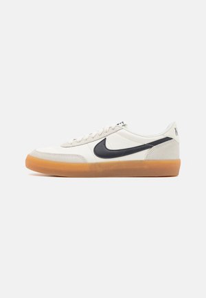 Valkoinen matalakärkinen tennari, jossa musta Nike-logo, kumipohja ja nauhakiinnitys, esitetty sivuprofiilissa yksinkertaisella taustalla.