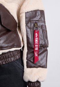 Giacca di pelle marrone con rivestimento in shearling crema, polsini a coste e un'etichetta rossa di stoffa che recita "RIMUOVERE PRIMA DEL VOLO".