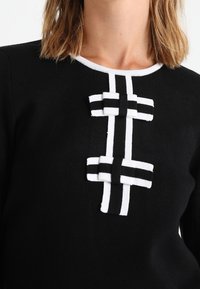 Černý svetr s bílými detaily má dvě dekorační černobílé pruhované detaily ve stylu stahovacích knoflíků na hrudi pod oblým výstřihem.