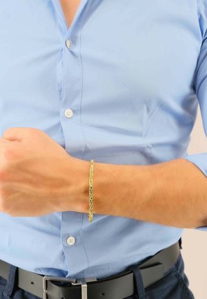Bracciale dorato con un motivo intricato, indossato su un polso. Chi lo indossa ha una camicia azzurro chiaro con bottoni e una cintura nera.