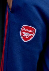 Modré tepláky se červenými pruhy, které mají logo Arsenal FC v červené a bílé barvě. Logo obsahuje kanón a kulaté designové detaily.