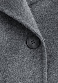 Pilkas vilnos audinys su minkštu, raštuotu paviršiumi. Apatinėje dalyje yra apvalus juodas mygtukas su keturiomis skylutėmis, kuris yra pritvirtintas prie apykaklės.