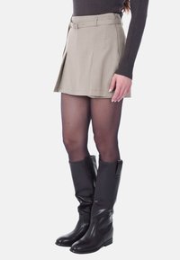 Gonna plissettata beige con vita con cintura, abbinata a stivali neri al ginocchio e collant neri trasparenti, che mostrano un design sartoriale.