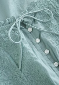 Vêtement bleu clair avec des motifs floraux brodés, quatre boutons blancs et un nœud en tissu noué près de l'encolure.
