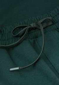 Donkergroene joggerbroek met een tailleband met trekkoord en een textuurkoord met metalen aglet. De stof lijkt zacht en glad te zijn.