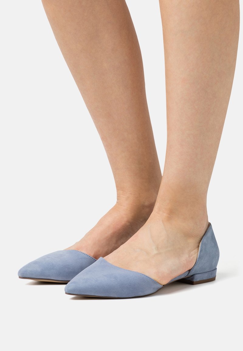 Femme portant des chaussures plates pointues bleu clair à petit talon, pieds nus, debout sur un fond blanc.