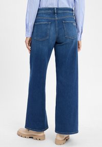 Weite blaue Jeans aus Denim, mit hoher Taille, zwei Gesäßtaschen und einfacher Naht. Kombiniert mit beigen Plateauschuhen.