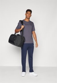 Schwarze Sporttasche, die von einer Person in einem grauen T-Shirt mit "Champion"-Logo, marineblauen Jogginghosen und weißen Turnschuhen gehalten wird. Schlichtes Design und Materialien.