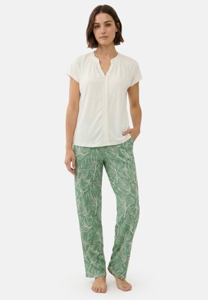 Vrouw staat op blote voeten, draagt een witte blouse met korte mouwen en een groene broek met wit bladmotief, één hand in de zak, neutrale achtergrond.