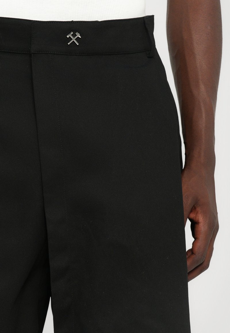 Svarte skræddersyede bukser med flad front, subtil tekstur og en metallisk logo accent med krydsede værktøjer i taljen.