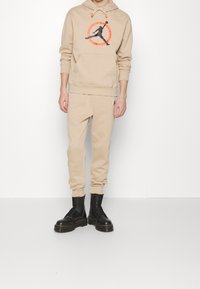 Beige Kapuzenpullover mit einem schwarzen Grafiklogo und orangefarbenen Akzenten, kombiniert mit passenden beige Jogginghosen und schwarzen Schnürstiefeln.