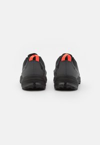 Czarne buty sportowe z teksturowaną cholewką, z grubą podeszwą i neonowo-pomarańczową pętelką na pięcie. Projekt jest smukły z minimalnymi akcentami.
