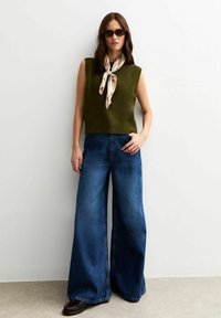 Olijfgroen gebreid mouwloos topje gecombineerd met hoge taille, wijdvallende blauwe denim jeans. Een gestructureerde crèmekleurige sjaal en donkere zonnebril maken de look compleet.