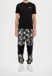 Black cotton t-shirt with gold logo; multi-colored camouflage joggers with black fabric accents; white sneakers. Simple, athletic fit.