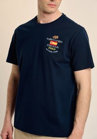 Camiseta de algodón azul marino con logotipos bordados, que incluyen un caballo y la bandera española, con mangas cortas y corte relajado.
