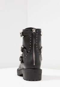 Stivale alla caviglia in pelle nera con cerniere argentate, fibbie, borchie e decorazioni in perle su una superficie e sfondo bianco.