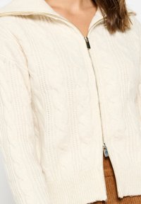 Kabelinis šviesiai kreminės spalvos megztinis su užtrauktuku priekyje ir plačiu apykakle. Yra tekstūruoti raštai ir gyslotas rankovės kraštas, demonstruojantis minkštą mezginį.