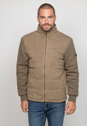 Homme aux cheveux courts portant une veste matelassée brune avec des manches texturées et un jean bleu, debout devant un fond clair uni.