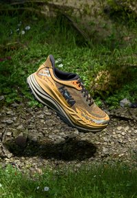 Scarpa da corsa Hoka con tomaia a motivo giallo e grigio, lacci neri e suola imbottita, sospesa sopra un ambiente naturale di erba e pietre.