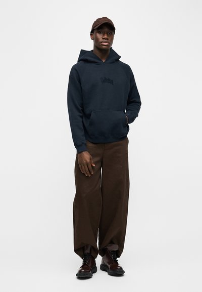 Άνδρας που φοράει σκούρο μπλε φούτερ με κουκούλα, καφέ παντελόνι με φαρδιά πόδια, σκούρα καφέ μπότες και καφέ καπέλο, όρθιος μπροστά σε απλό λευκό φόντο.