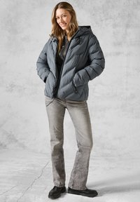 Doudoune grise avec capuche, présentant des détails cousus et une fermeture éclair. Associée à un jean gris clair et des chaussures noires.