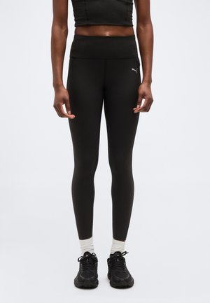 Vrouw draagt zwarte hoge taille Puma-legging met een cropped zwart topje, witte sokken en zwarte sportschoenen, staand tegen een effen achtergrond.