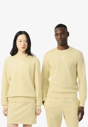 Twee personen dragen bijpassende lichtgele sweatshirts. De outfit van de vrouw omvat een bijpassende lichtgele rok, beide met een minimaal logo.