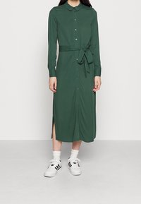 Kobieta w zielonej, midi sukience z długim rękawem i zapięciem na guziki, z paskiem w talii, w połączeniu z białymi skarpetkami i białymi trampkami Adidas w czarne paski.