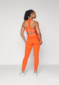 Oranžinis sportinis kostiumas su nugaros kirpimu ir logotipo detale, taip pat aukšto liemens leggingai. Lygus tekstūra, prigludęs dizainas ir kulkšnies ilgio.