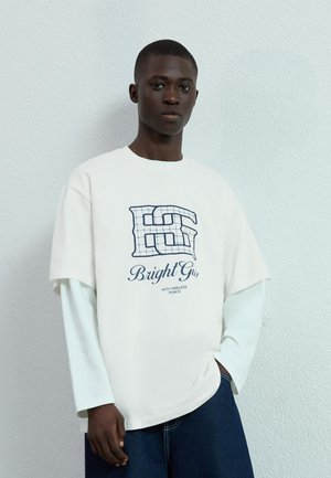 Un tricou alb supradimensionat cu un logo în carouri albastre și textul "BrightGu", purtat peste un top cu mânecă lungă de culoare bleu deschis, asortat cu blugi negri.