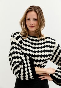 Schwarz-weißer Pullover mit einem geometrischen Muster, weiten Ärmeln und einer strukturierten Oberfläche. Der Halsausschnitt ist rund und das Material wirkt dick.