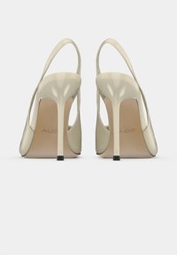 Sandales à talons hauts beiges avec une tige transparente, un dos ouvert et de fines lanières autour de la cheville. Elles présentent un talon effilé et un logo discret sur la semelle intérieure.