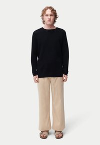 Pull noir côtelé, pantalon ample en lin beige et sandales marron. Le modèle se tient devant un fond neutre, les bras le long du corps.
