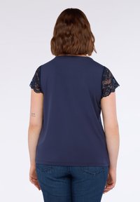 Donna con capelli castani a spalle indossa una maglia blu navy con maniche corte in pizzo e jeans blu, mostrata di spalle su uno sfondo neutro.