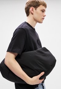 Melnas mīkstas kabatas soma, kas turēta zemā rokā, izgatavota no maigas auduma ar gludu faktūru. Soma ir taisnstūrveida un izskatās viegla.