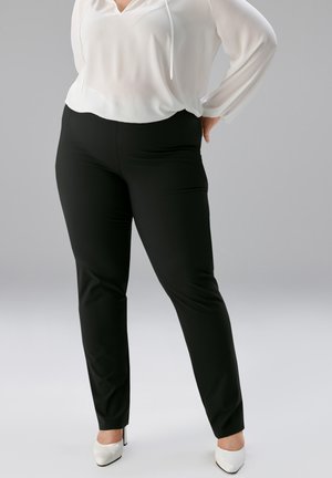 Frau trägt schwarze eng anliegende Hose, weiße Bluse mit Kordeln und weiße High Heels, steht mit einer Hand in der Hüfte.