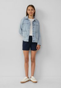 Hellblaues Denim-Jackett mit großen Knöpfen, weißem T-Shirt, strukturierten dunkelblauen Shorts und weißen Sneakers mit braunen Sohlen.