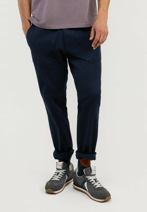 Hombre con pantalones azul oscuro con dobladillos remangados, calcetines marrones con dibujos y zapatillas grises, blancas y azules de pie frente a un fondo blanco.
