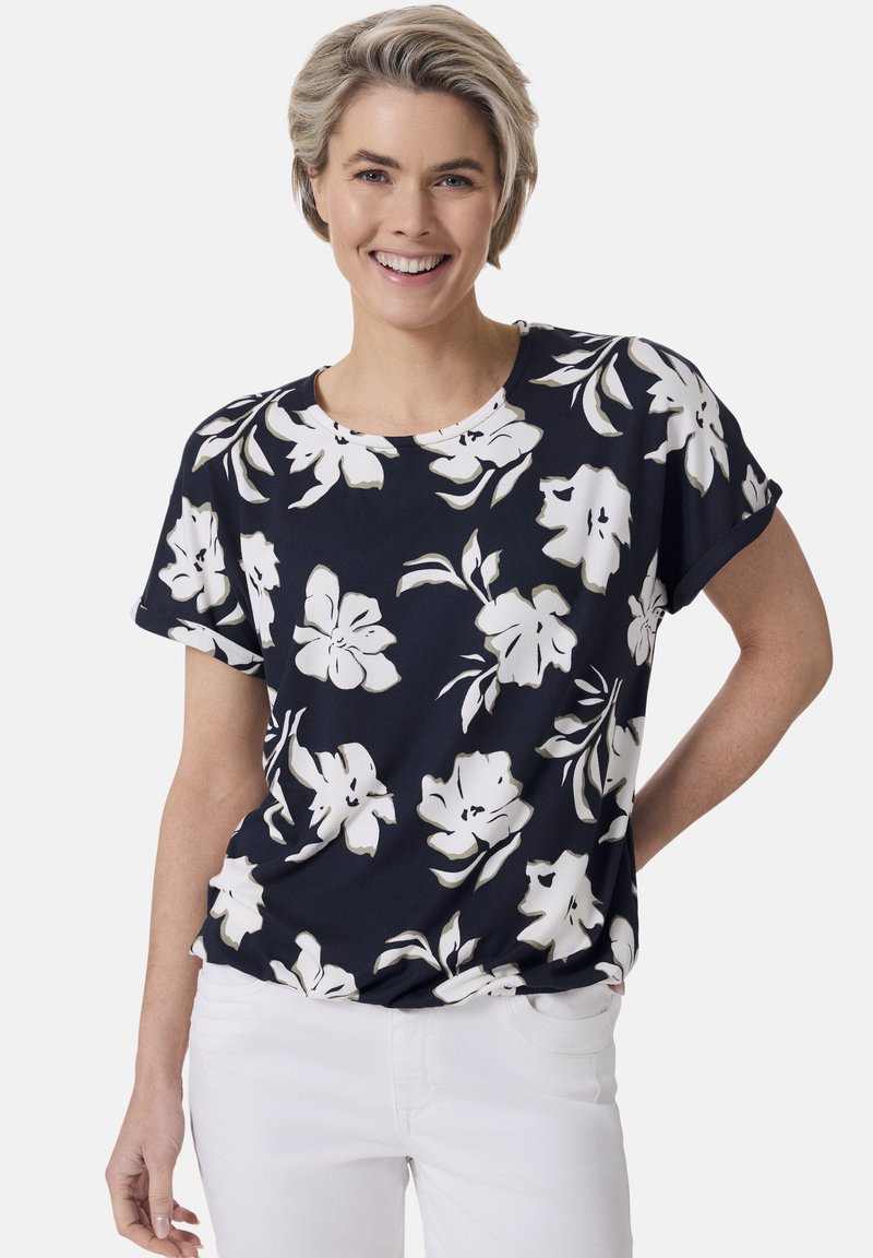 Marineblaues floral bedrucktes T-Shirt mit kurzen Ärmeln, weißen und beigen Blumenmustern, runder Ausschnitt, weicher Stoff, kombiniert mit weißen Hosen.