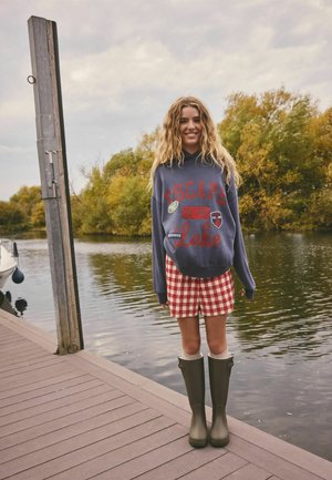 Jauna moteris šypsosi ant medinės liepto ežero pakrantėje, dėvi tamsiai mėlyną džemperį su gobtuvu, raudoną languotą sijoną, žalius iki kelių siekiančius batus, fone – rudens medžiai.