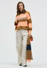 Striped sweater in orange, pink, and brown with white text "Senses." Paired with loose cream pants and a matching scarf in similar colors.