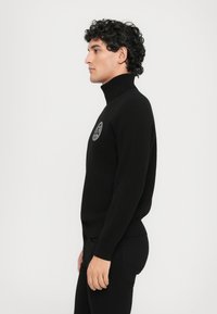 Μαύρο πλεκτό ζιβάγκο με ριγέ υφή και κυκλικό λογότυπο στη στήθος. Στενή γραμμή, φορεμένο με σκούρα παντελόνια.